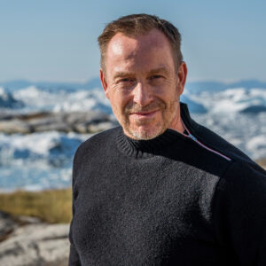 Portræt af Adam Price. Man i blå sweater kigger ind i kameraet. Hav og is i baggrunden.